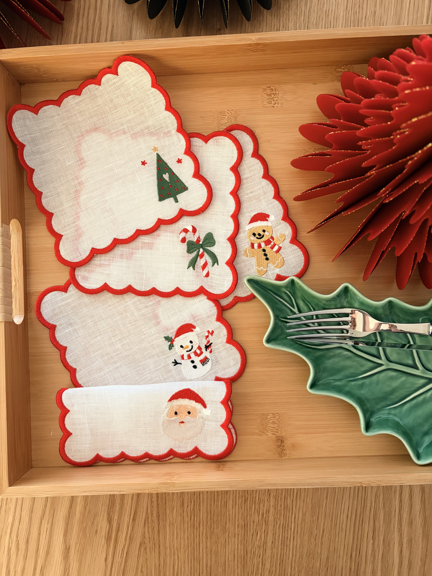 Guardanapos de Natal de tecido branco com bordas vermelhas e desenhos natalícios sobre tabuleiro de madeira.