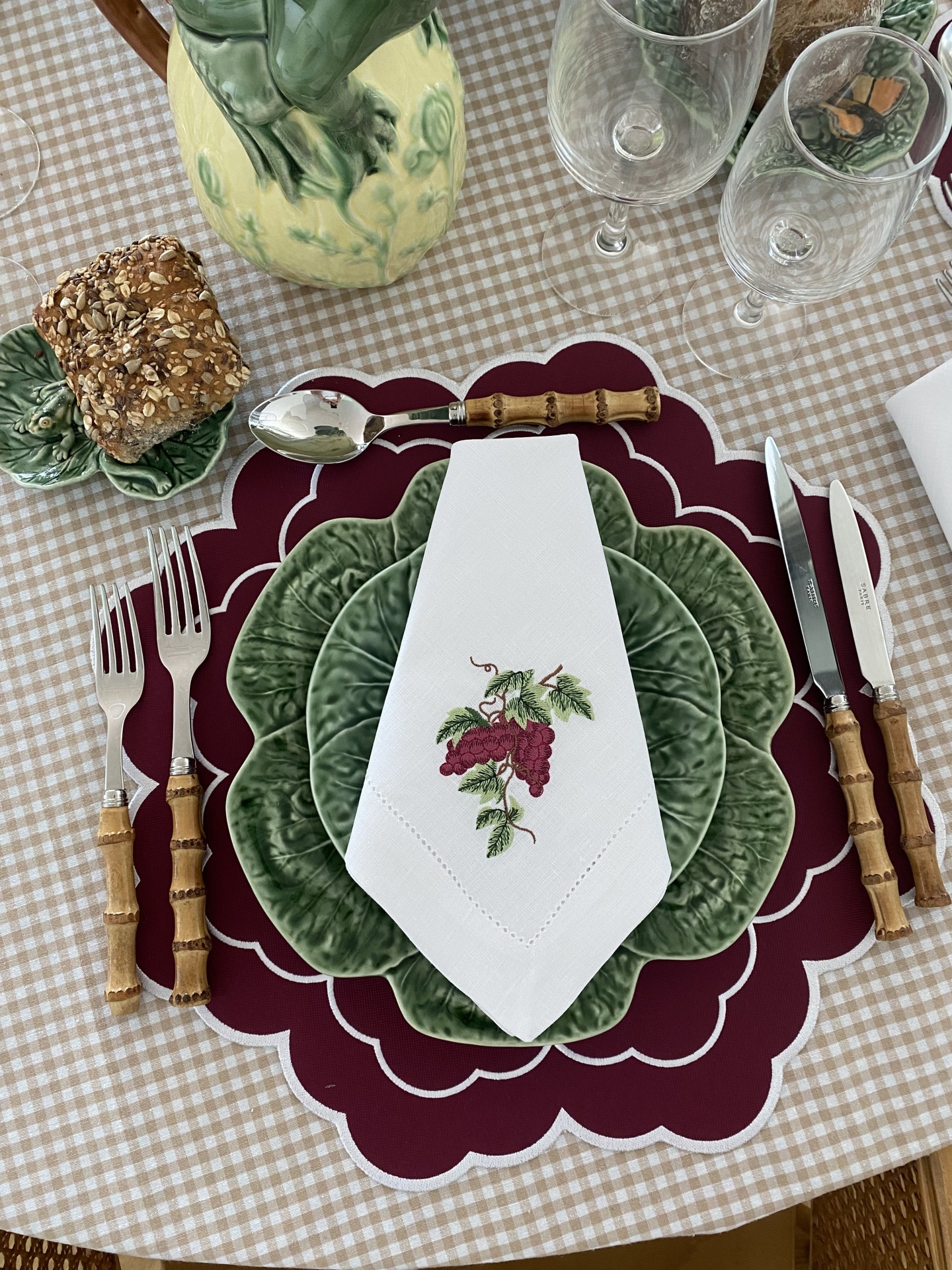 Mesa posta com pratos verdes em forma de folha, talheres com cabo tipo bambu, guardanapo branco com bordado de uvas e pão integral num pires verde