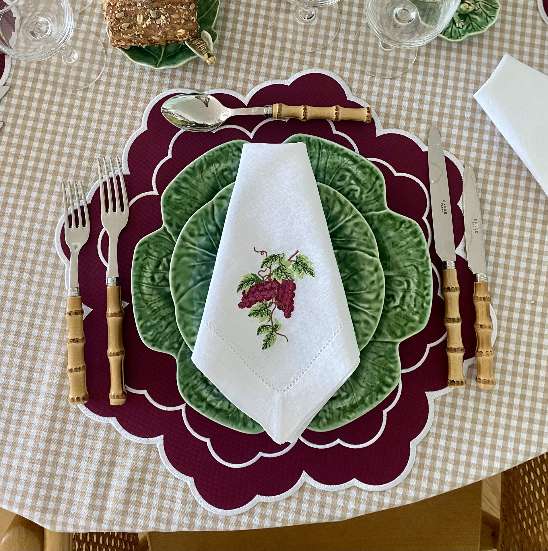 Mesa posta com pratos verdes, guardanapo branco bordado com uvas, talheres com cabo de bambu, base de prato vinco vermelho e toalha xadrez bege