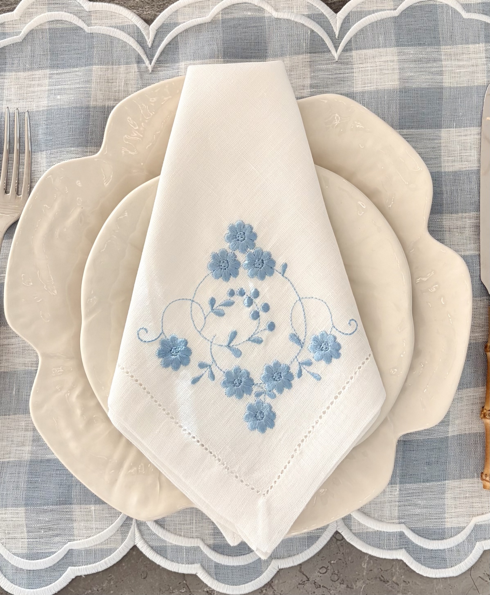Guardanapo branco bordado com flores azuis sobre pratos brancos em toalha xadrez azul e branca.