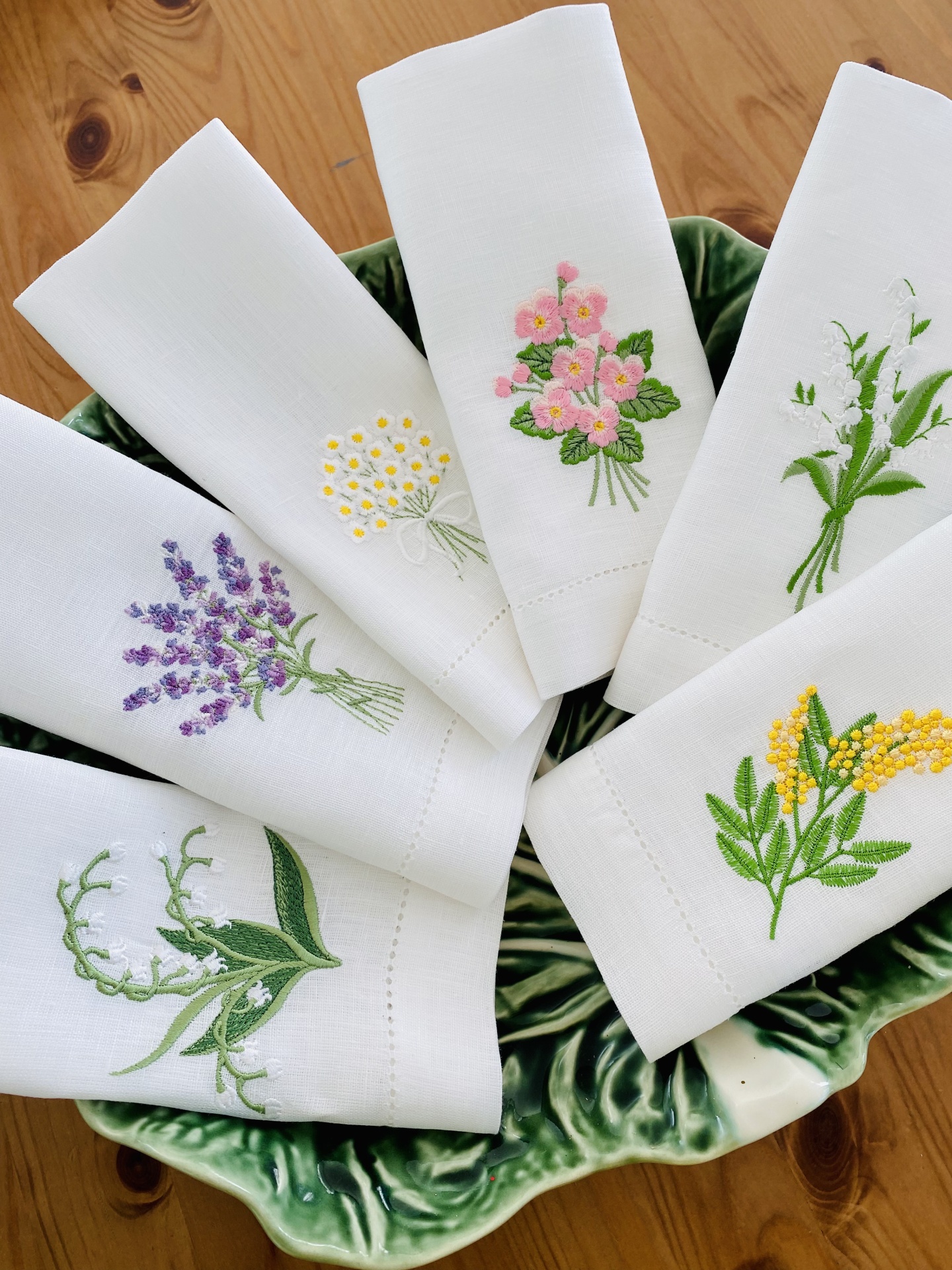 Seis guardanapos brancos bordados com flores coloridas numa travessa verde em mesa de madeira