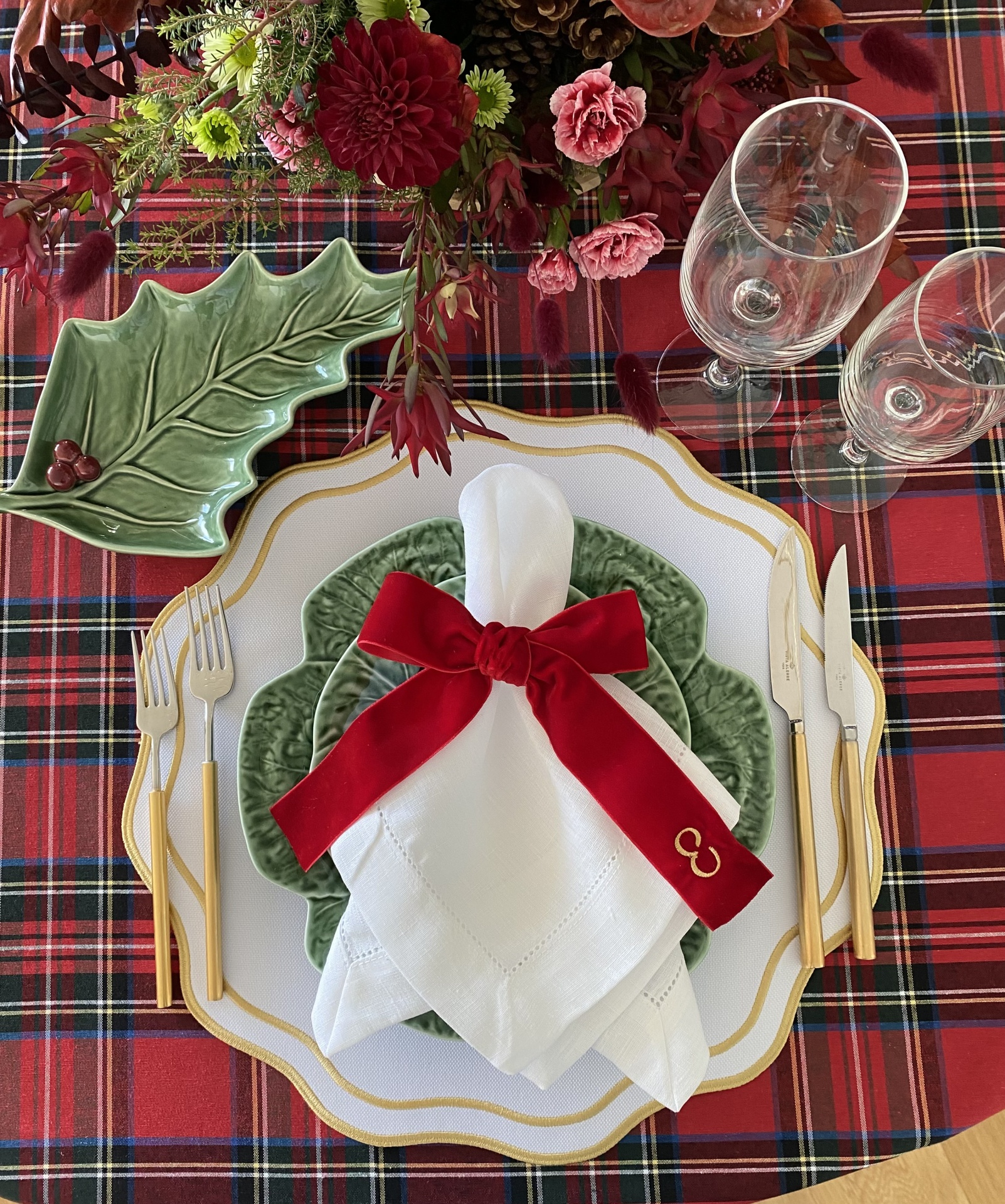 Mesa posta com pratos brancos e verdes, talheres dourados e arranjo floral sobre toalha xadrez vermelha