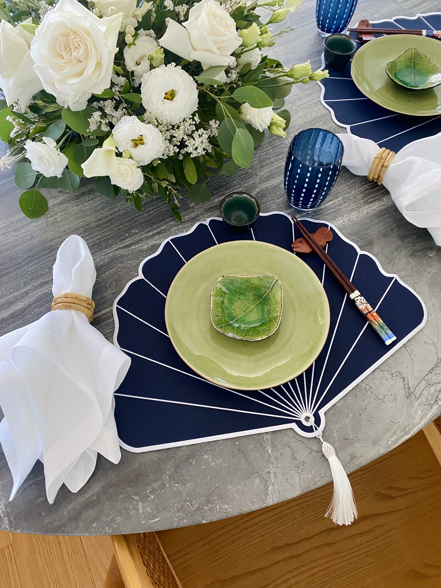 Mesa posta de jantar com prato verde, passadeira azul em forma de leque, copo azul e flores brancas