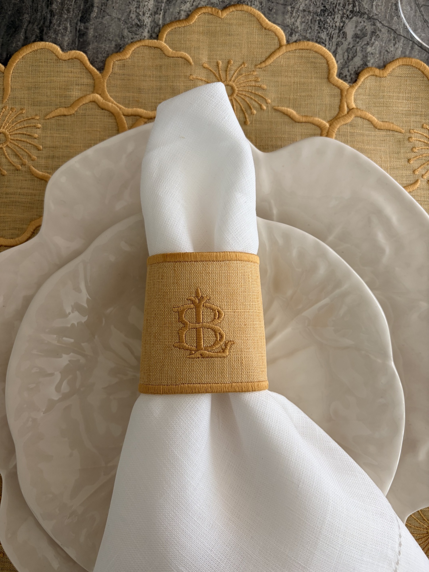 Conjunto de loiça com pratos cerâmicos creme, guardanapo branco com argola bordada amarela e toalha floral amarela.