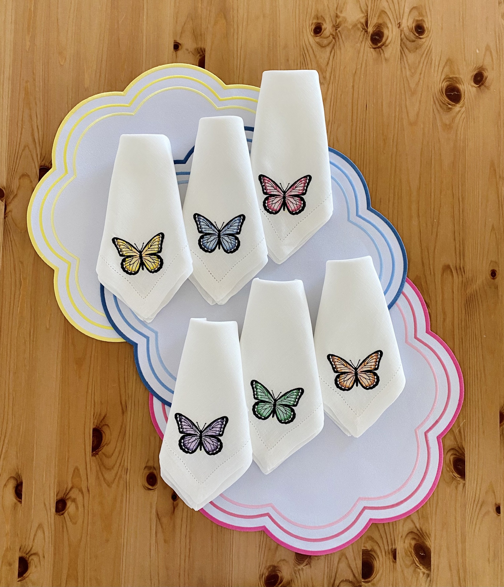 Guardanapos brancos bordados com borboletas coloridas e bases de pratos com bordas coloridas sobre mesa madeira