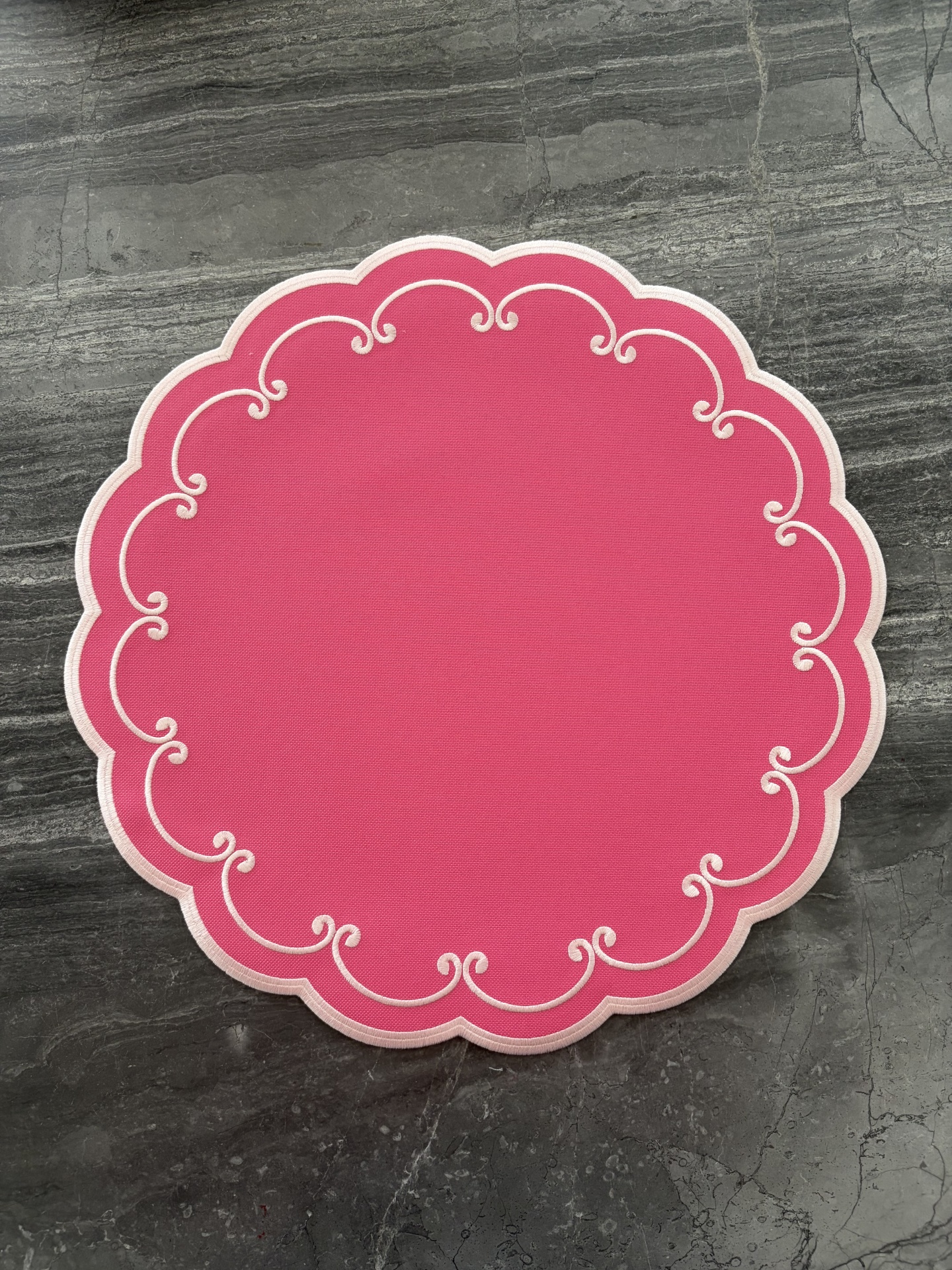 Individual redondo de tecido vermelho com bordado branco em arabescos sobre fundo de mármore cinza.