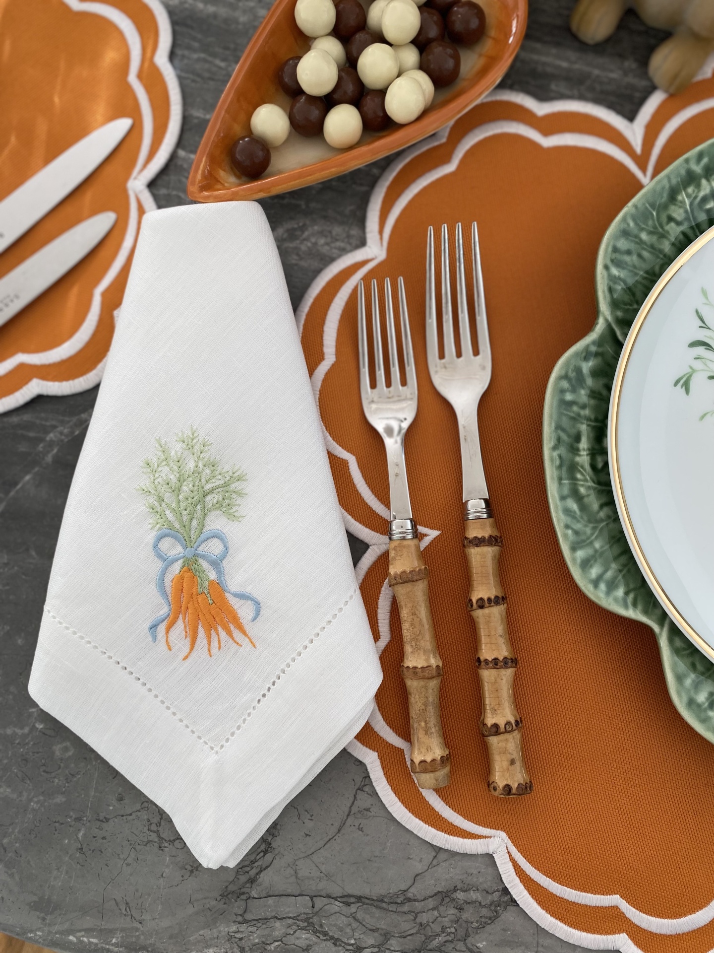 Guardanapo branco bordado com cenouras, dois garfos com cabo de madeira, individuais laranja com borda branca e prato verde sobre mesa cinza