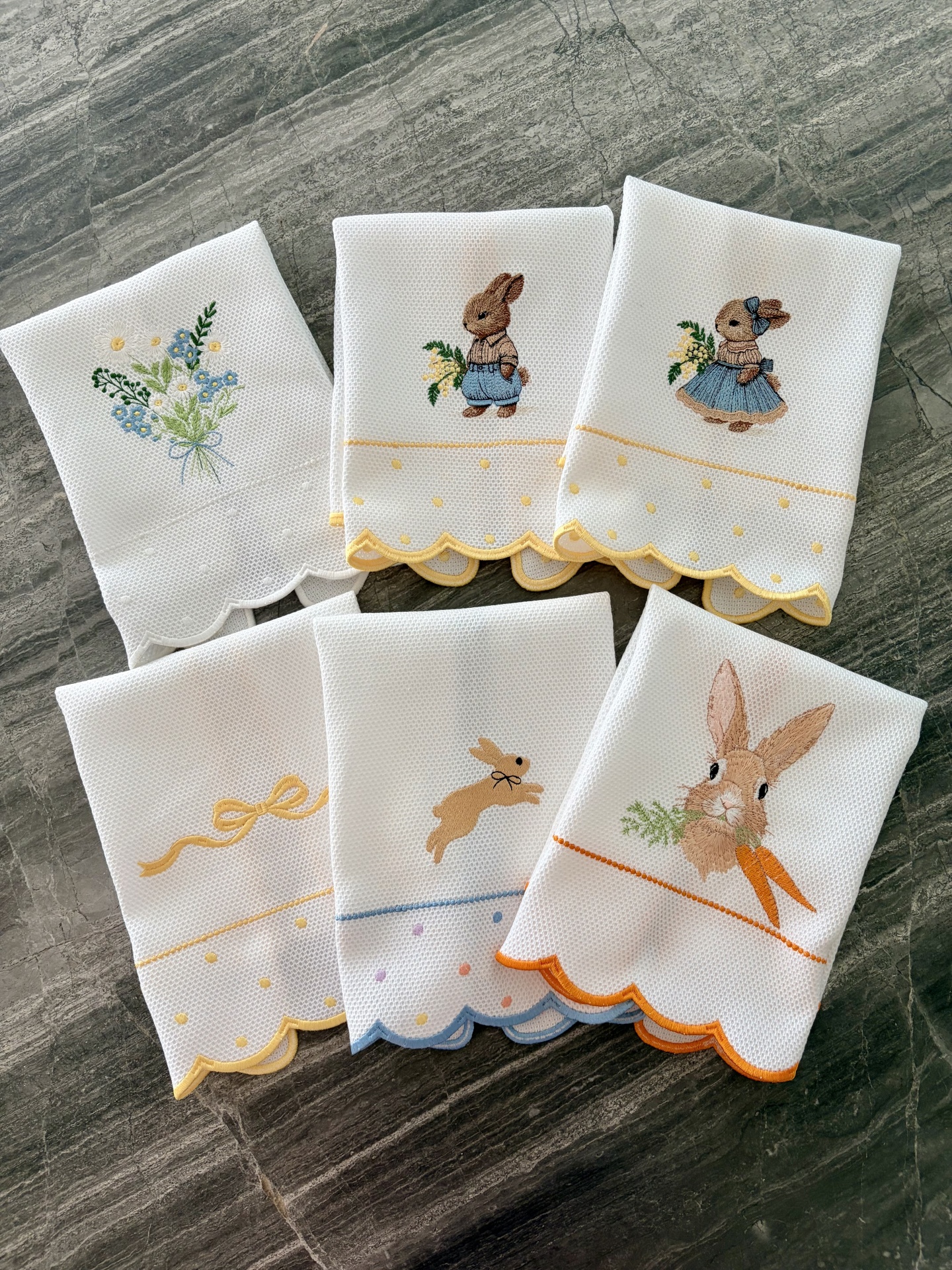 Paninhos de cozinha brancos com bordados de coelhos, flores e laços e bordas coloridas sobre superfície cinzenta.