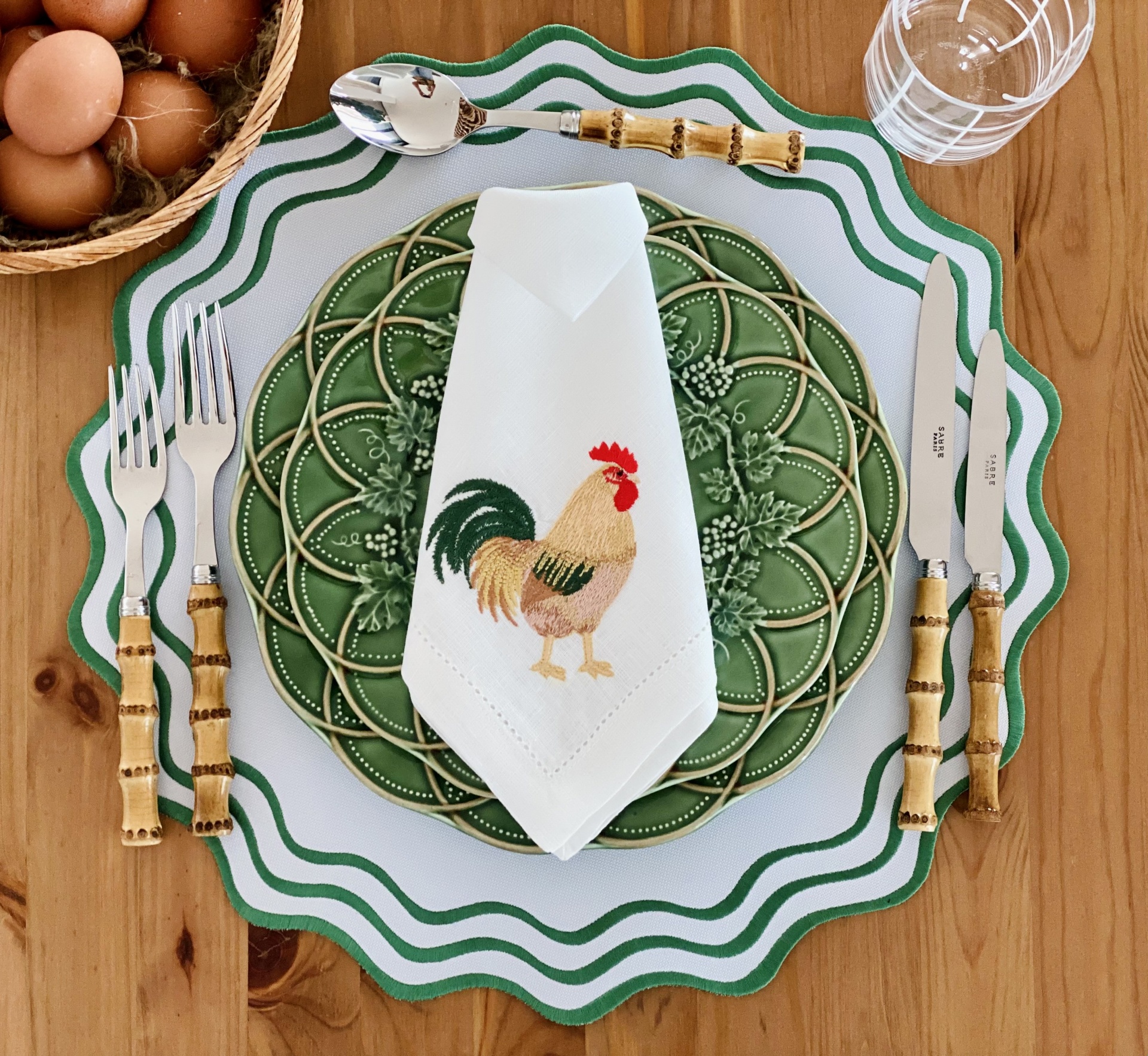 Mesa posta com pratos brancos e verdes, guardanapo bordado, talheres com cabo de bambu, ovos numa cesta e copo sobre mesa de madeira.