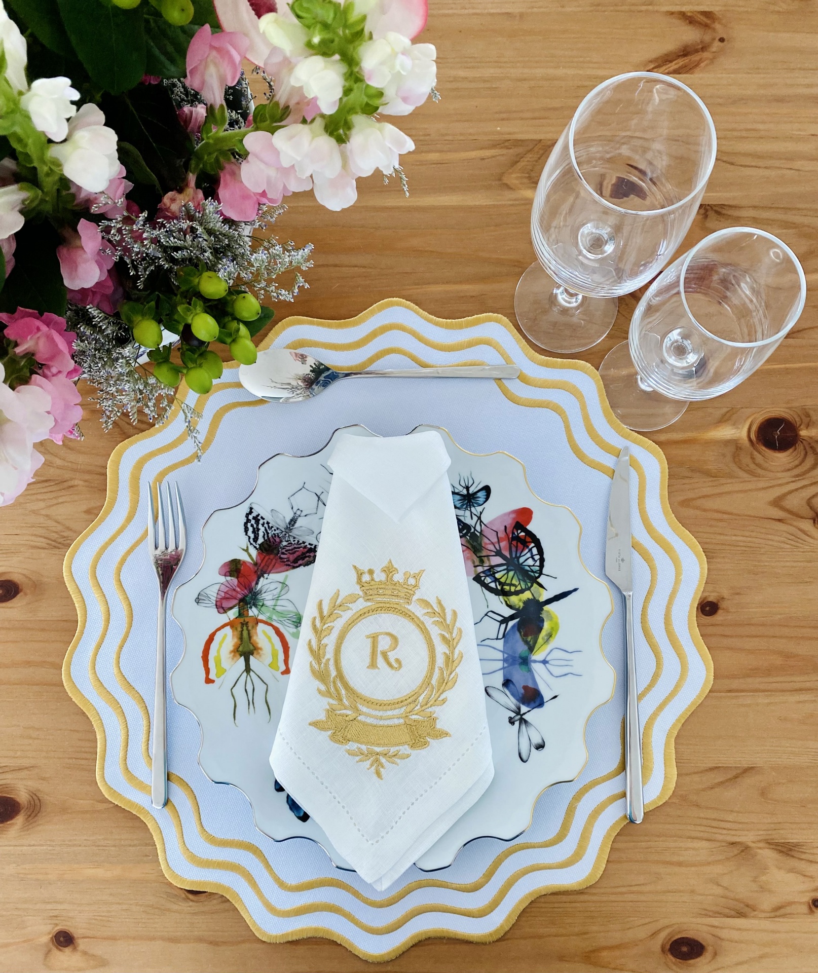 Conjunto de jantar com pratos decorativos, guardanapo com monograma e dois copos em mesa de madeira