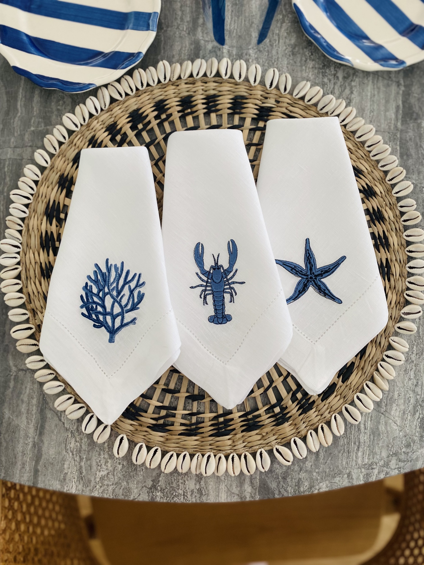 Três guardanapos brancos bordados com motivos marinhos azuis sobre prato de verga e búzios