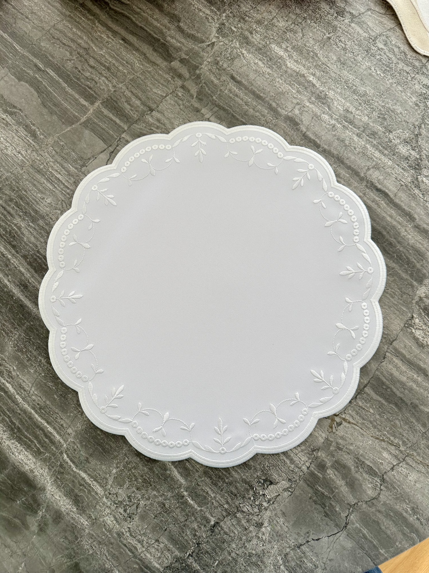 Toalha de mesa redonda branca com bordado de folhas e círculos em superfície de pedra cinza.