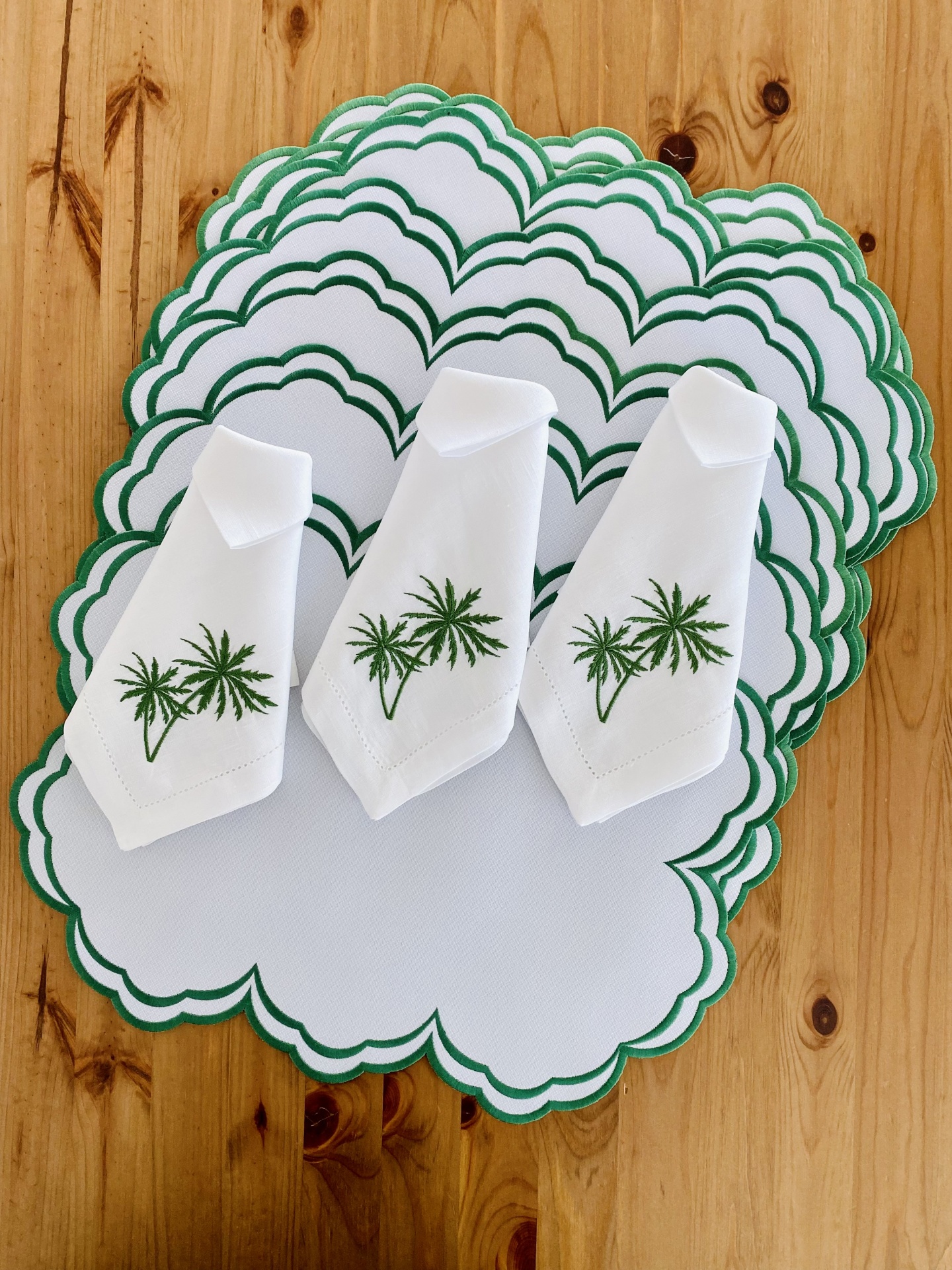 Jogo americano e guardanapos brancos com bordado de palmeira verde e bordas onduladas verdes sobre mesa de madeira.