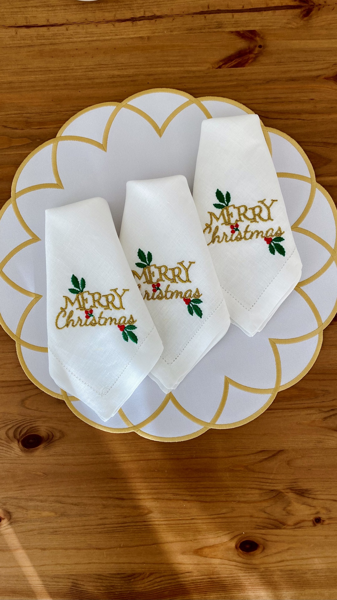 Três guardanapos brancos bordados com 'Merry Christmas' em oro e verde, sobre um jogo americano branco com linhas douradas, numa mesa de madeira.