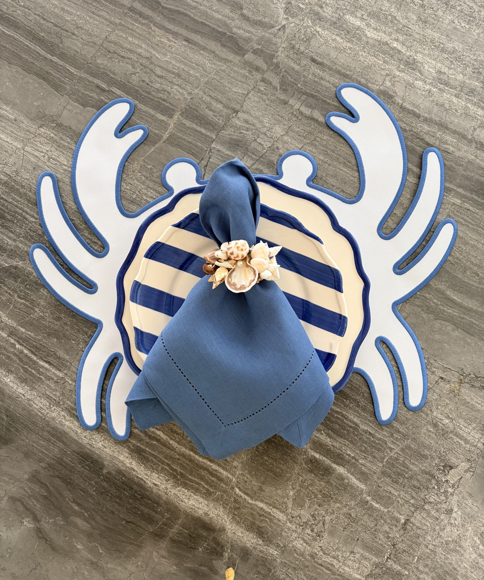 Conjunto de loiça com prato às riscas azuis e creme, guardanapo azul com argola de conchas, base em padrão coral azul e branco, superfície cinza