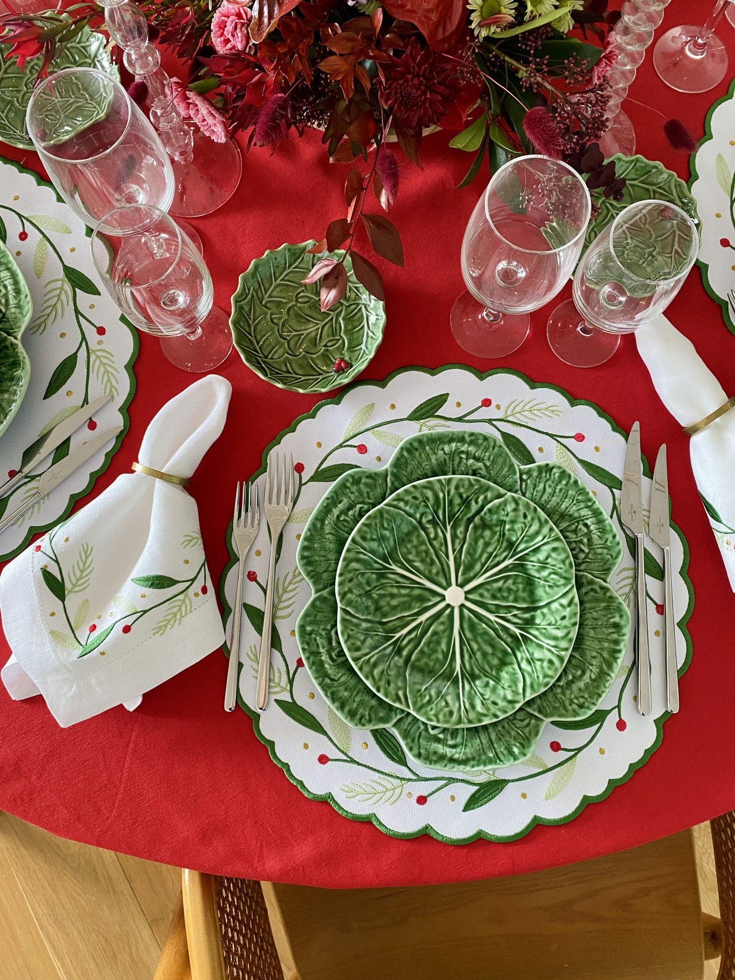 Mesa posta com pratos verdes em formato de folhas, guardanapos com padrão de folhas e bagas, talheres e copos sobre toalha vermelha