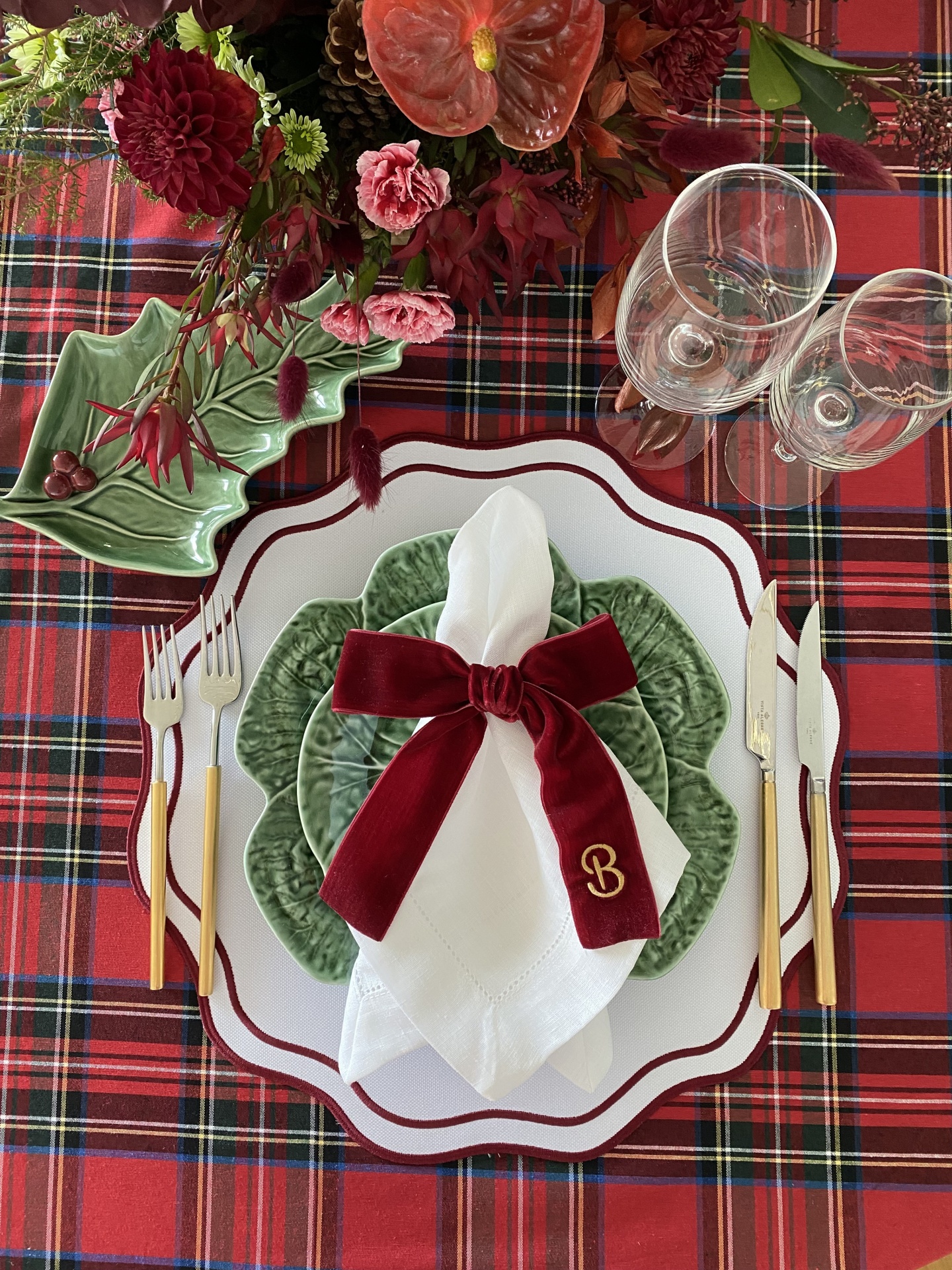 Mesa posta com conjunto de jantar em pratos brancos e verdes sobre toalha xadrez e arranjo floral vermelho