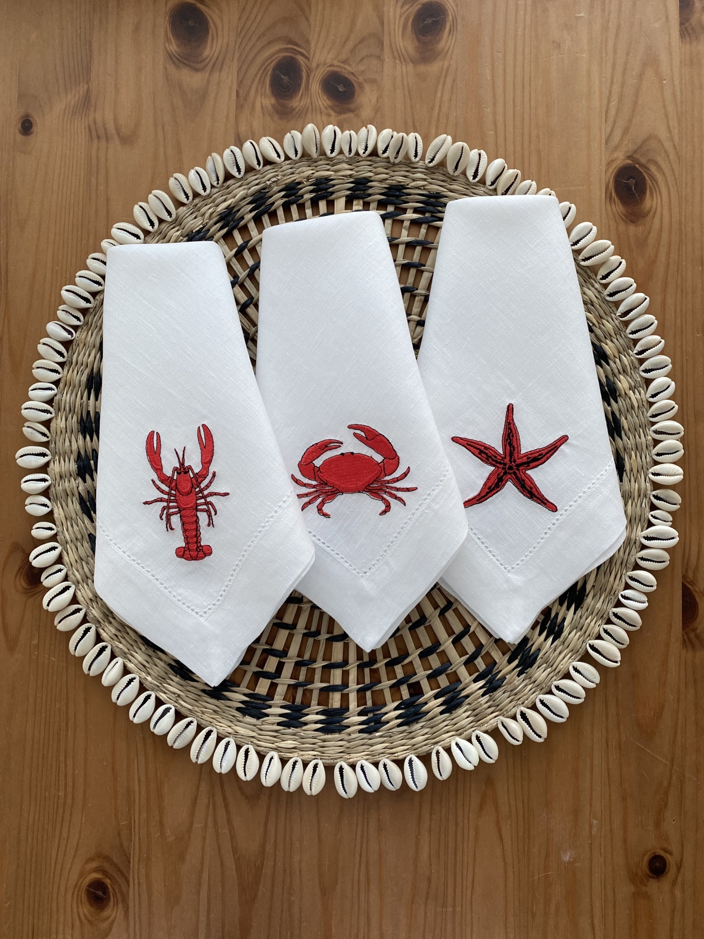 Três guardanapos brancos com bordados vermelhos de animais marinhos sobre prato trançado e conchas.