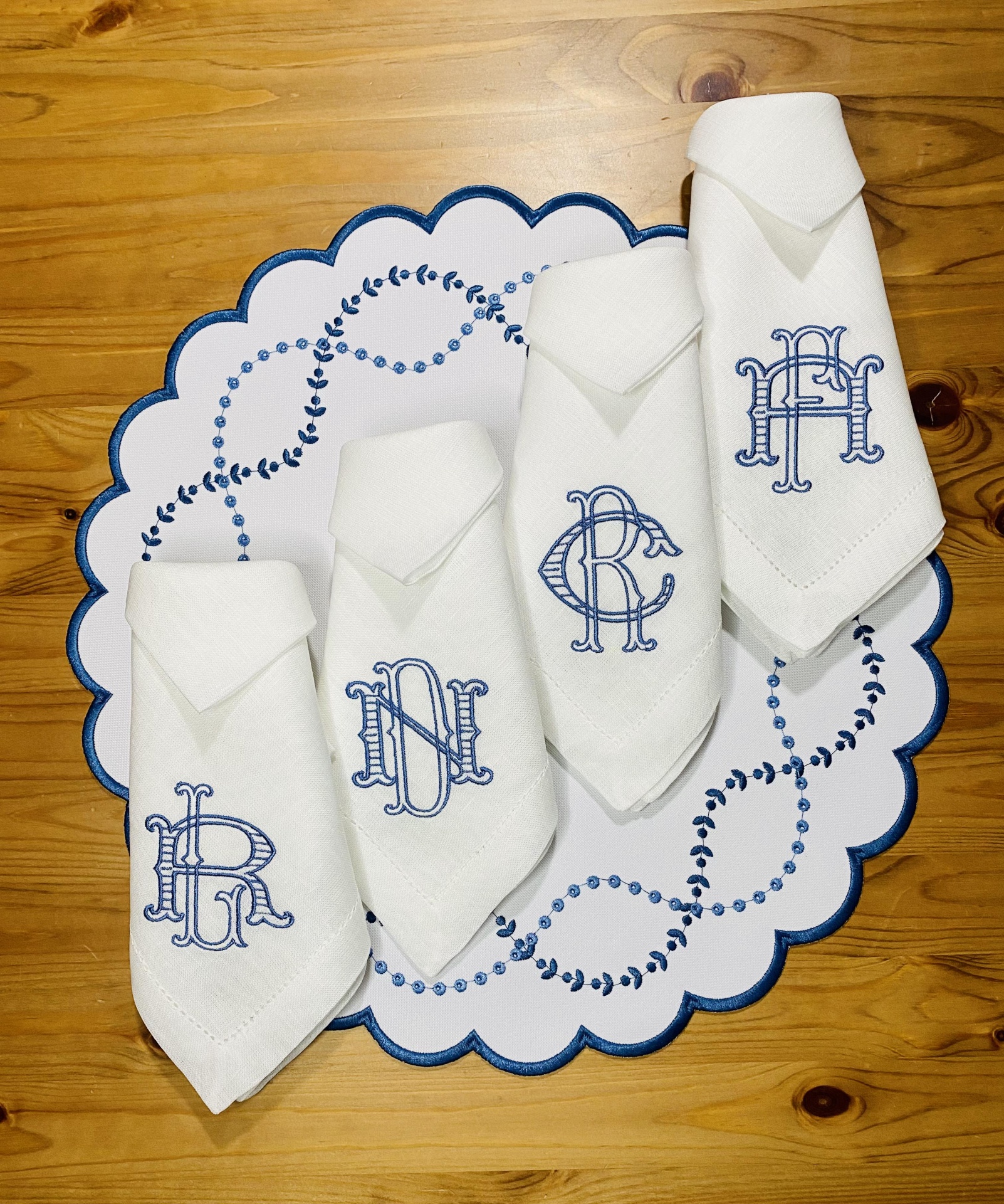 Guardanapos brancos bordados com letras azuis sobre sousplat branco com bordas azuis numa mesa de madeira