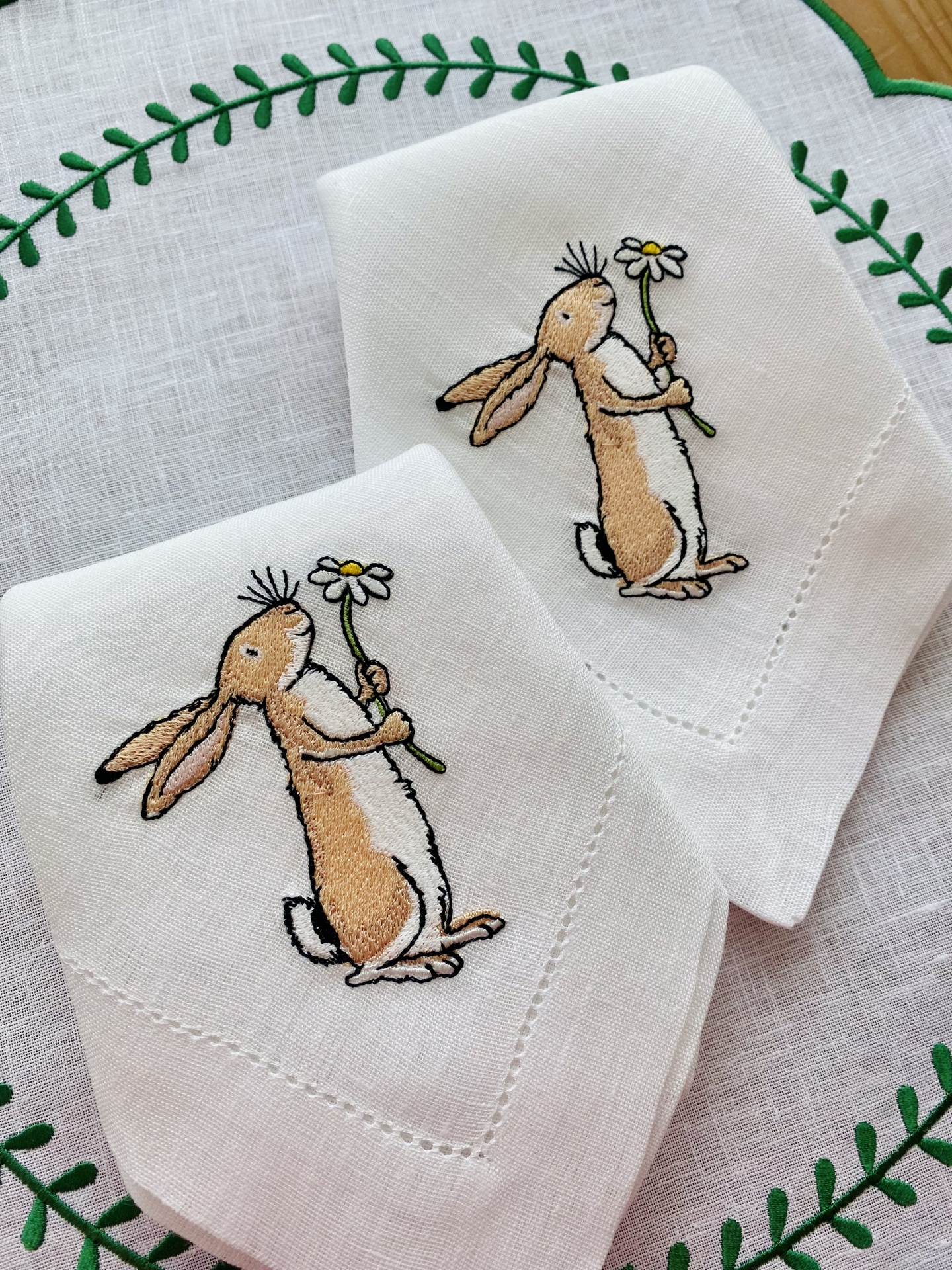 Guardanapos brancos bordados com coelhos segurando margaridas sobre tecido decorativo branco com folhas verdes.