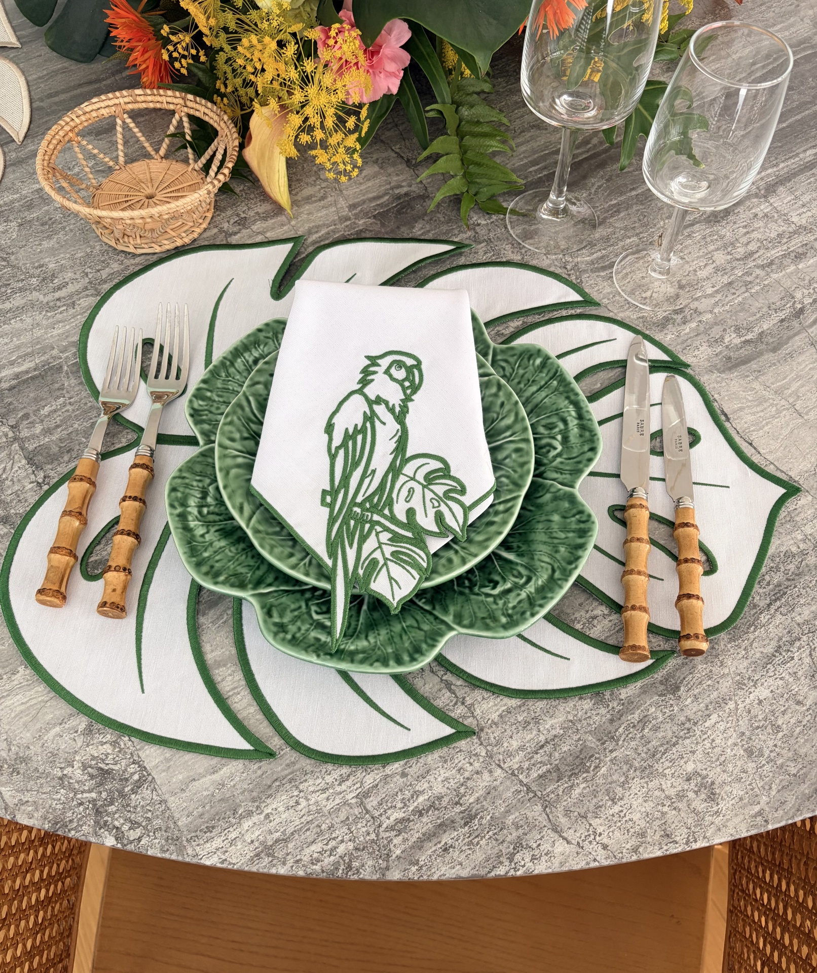 Mesa posta com prato verde em cerâmica, guardanapo branco com estampa de papagaio, talheres com cabo de madeira, copos transparentes, cesta de vime e arranjo floral