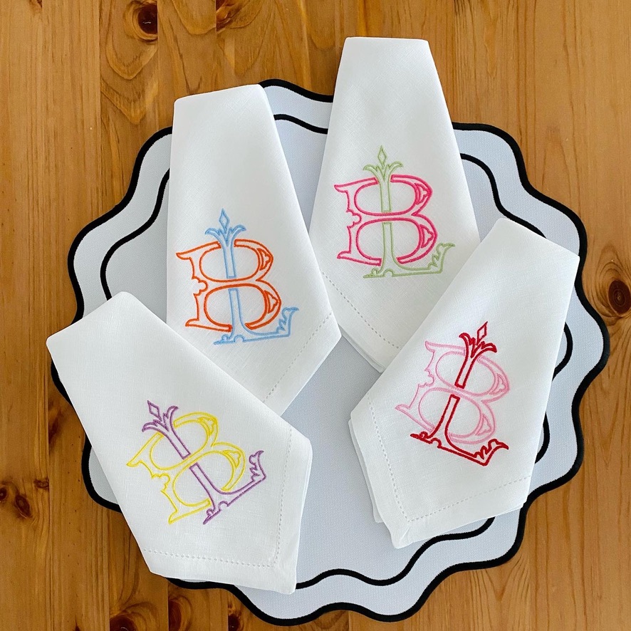 Conjunto de quatro guardanapos brancos com bordado colorido da letra B, sobre prato branco com borda preta, em mesa de madeira
