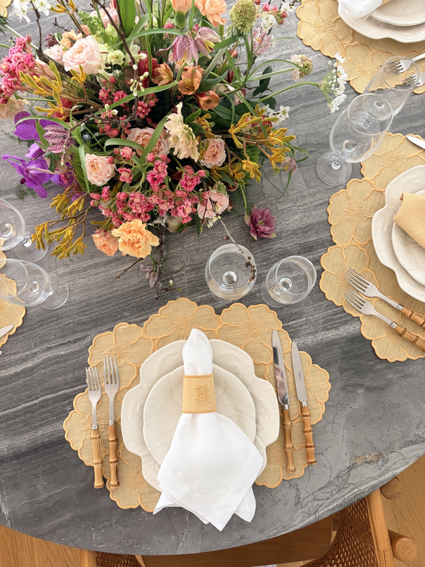 Mesa de jantar redonda em pedra cinza com louça branca, talheres com cabos em bambu, guardanapo branco, jogos americanos amarelos e arranjo de flores coloridas