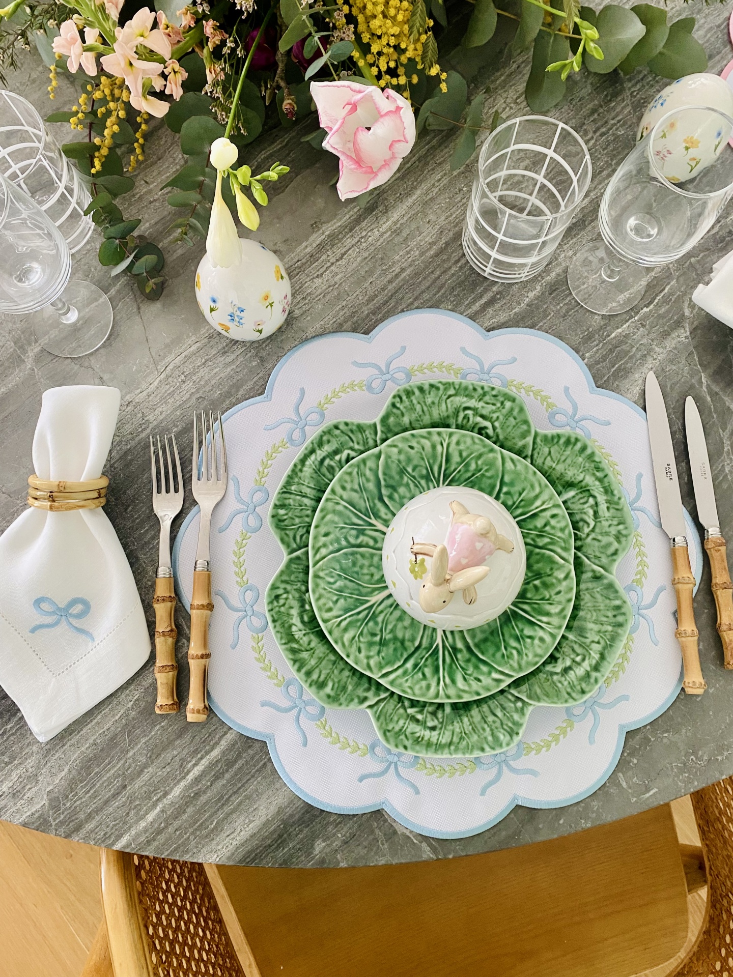 Mesa posta com pratos de cerâmica em forma de folhas verdes, talheres de cabo de madeira, guardanapo branco com bordado azul e anel dourado, copos transparentes e vaso branco com flores.