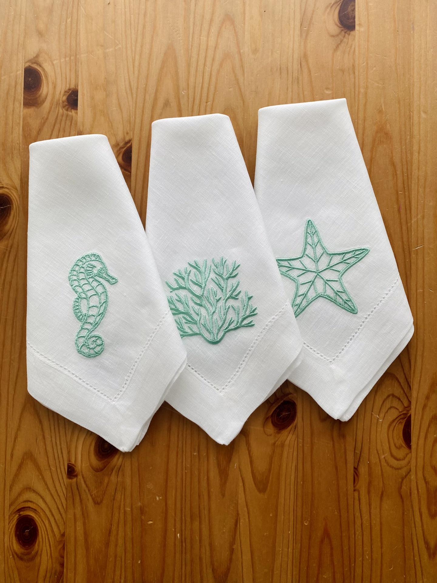 Três toalhas brancas com bordados verdes de mariscos sobre uma mesa de madeira