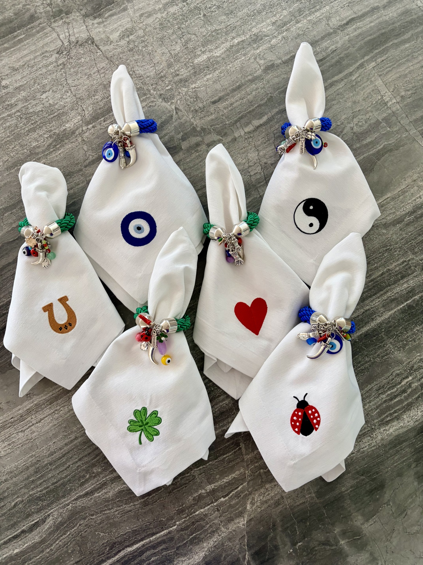 Conjunto de guardanapos brancos com bordados coloridos e anéis com pendentes sobre superfície cinza.
