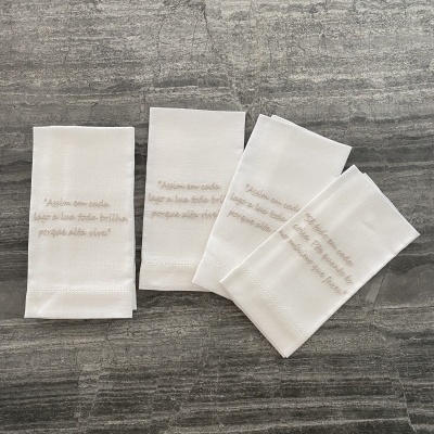 Quatro guardanapos de tecido branco com frase bordada em bege sobre superfície cinza