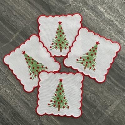 Quatro guardanapos brancos com borda vermelha e árvore de Natal bordada, sobre superfície cinza.