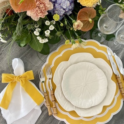 Conjunto de loiça branca com pratos amarelos, talheres de cabo madeira, guardanapo com laço amarelo e flores coloridas