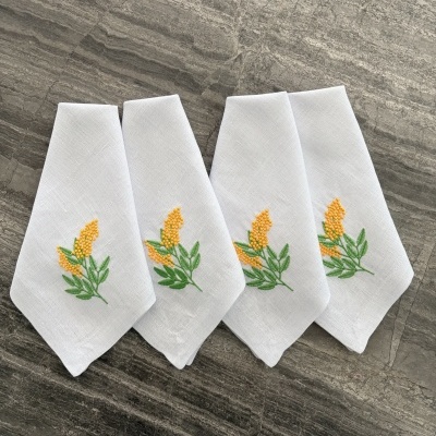 Quatro guardanapos brancos com bordado de flores amarelas e folhas verdes sobre pedra cinza.
