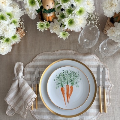 Conjunto de mesa com prato decorado com cenouras, talheres dourados, guardanapo às riscas e decoração com flores e coelho