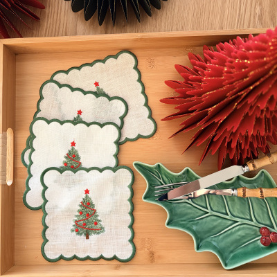 Toalhetes de Natal de linho branco com árvore verde, prato em forma de folha verde com frutas vermelhas e utensílios com cabo bambu numa bandeja de madeira clara.