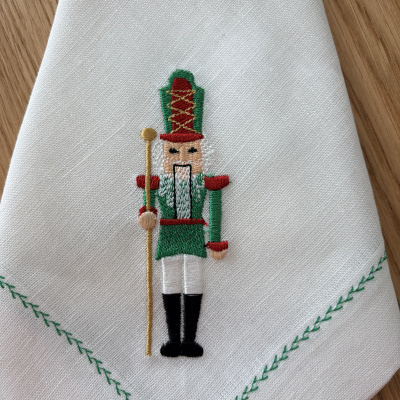 Guardanapo branco com bordado de soldado de brinquedo colorido e borda verde em padrão de folhas.