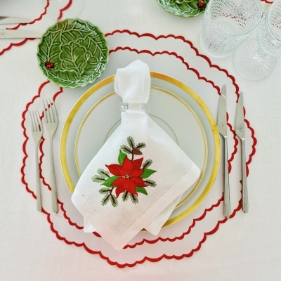 Conjunto de pratos brancos com borda dourada, guardanapo branco bordado em flor vermelha e folhas verdes, pratos verdes em relevo e copos de vidro sobre toalha branca com sousplats brancos e bordas vermelhas