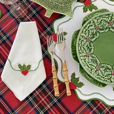 Mesa posta com pratos verdes decorados com folhas de azevinho, talheres com cabo de madeira, guardanapo branco com bordado de azevinho e toalha tartan.