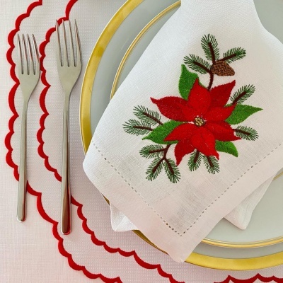 Guardanapo branco com bordado de flor de Natal vermelho e folhas verdes sobre prato branco com borda dourada e garfos, em toalha de mesa branca com borda vermelha