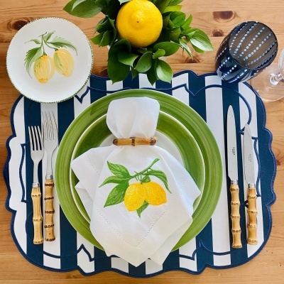 Mesa posta com prato verde, talheres de cabo castanho, prato com limões pintados, guardanapo branco com limões bordados e copo azul de bolinhas