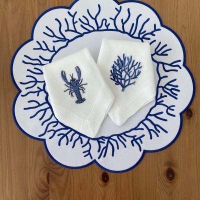 Guardanapos brancos bordados com desenhos azuis de coral e lagostim sobre descanso de prato branco e azul