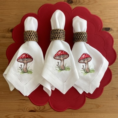 Três guardanapos brancos com cogumelos estampados e argolas em padrão castanho, sobre porta-copos vermelho, em mesa de madeira.