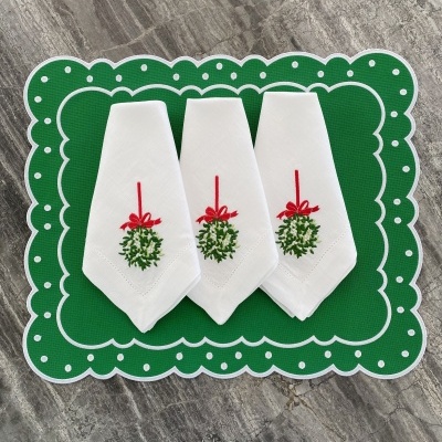 Três guardanapos brancos bordados com ramo verde e fita vermelha sobre tapete verde com pontos brancos