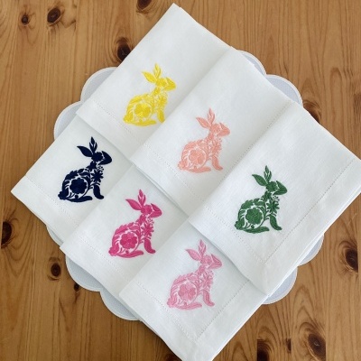 Cinco toalhas de mesa brancas com bordados coloridos de coelhos sobre prato branco e mesa de madeira.