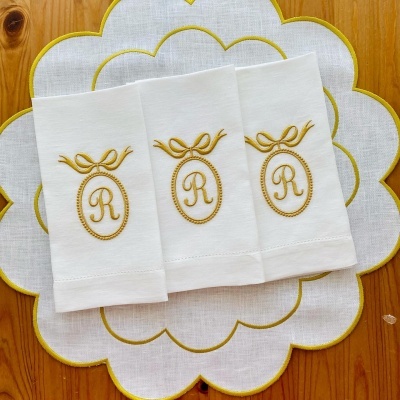 Guardanapos brancos bordados com a letra R dourada sobre um sousplat branco bordado a dourado