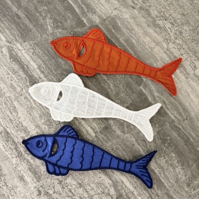 Três patches bordados em forma de peixe em cores laranja, branco e azul sobre pedra cinza.
