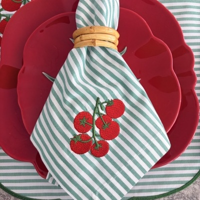 Conjunto de pratos vermelhos em forma de tomate com guardanapo às riscas verde e branco com bordado de tomates e argola de madeira.