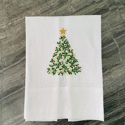 Guardanapo branco com árvore de Natal bordada em verde, vermelho e dourado sobre superfície de pedra cinzenta.