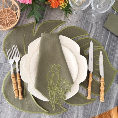 Mesa posta com toalha cinza, placemats verdes folhas, talheres bambu, pratos brancos, guardanapo verde bordado, copos vidro, flores coloridas.